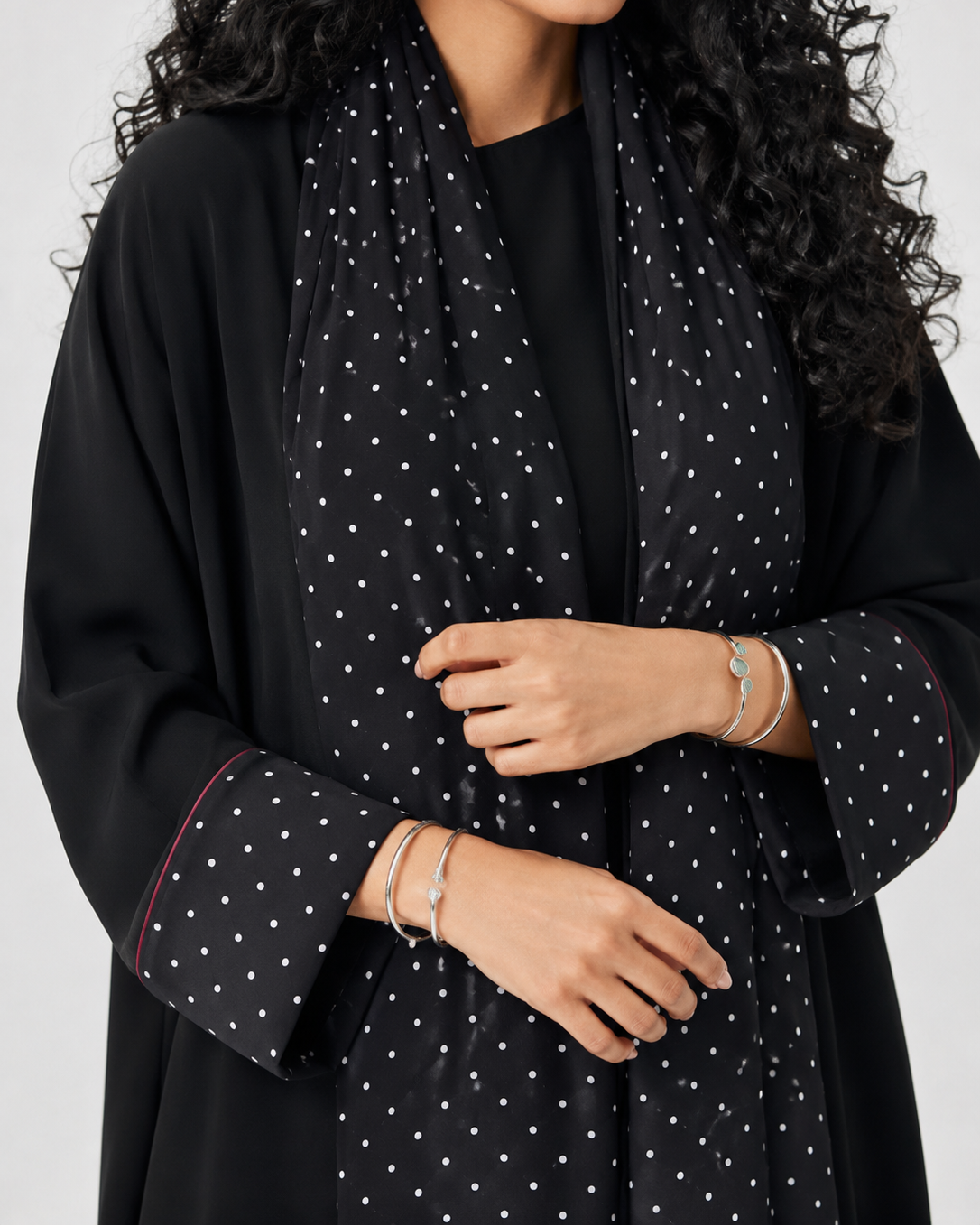 A close-up of a woman wearing a black long-sleeve abaya or robe with a white polka-dot patterned scarf, shown against a plain light gray background.