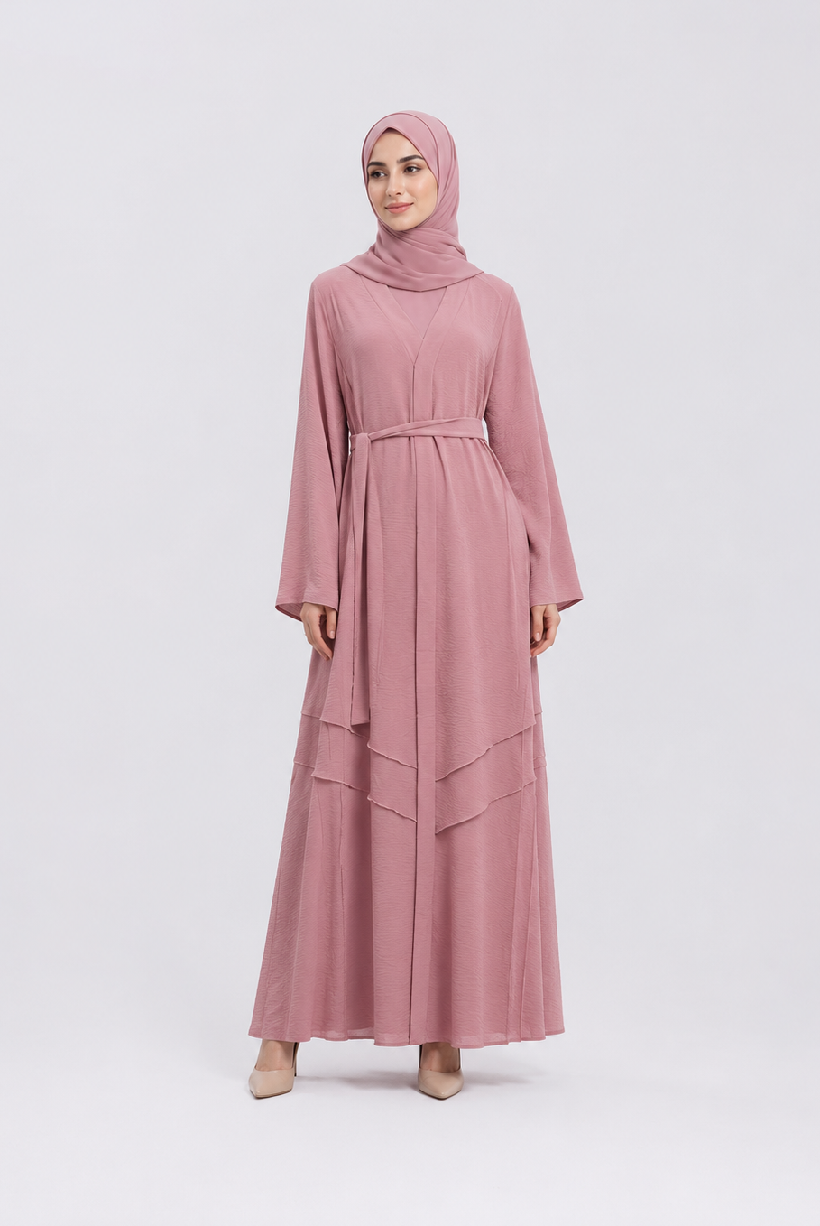 A woman modeling a dusty pink long-sleeved maxi abaya with a matching hijab, standing against a light gray studio background.