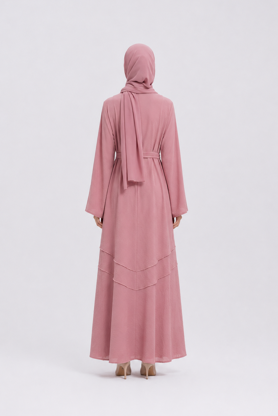 A woman viewed from the back wearing a long dusty pink abaya with a matching headscarf and a thin waist tie, standing against a light gray studio background.
