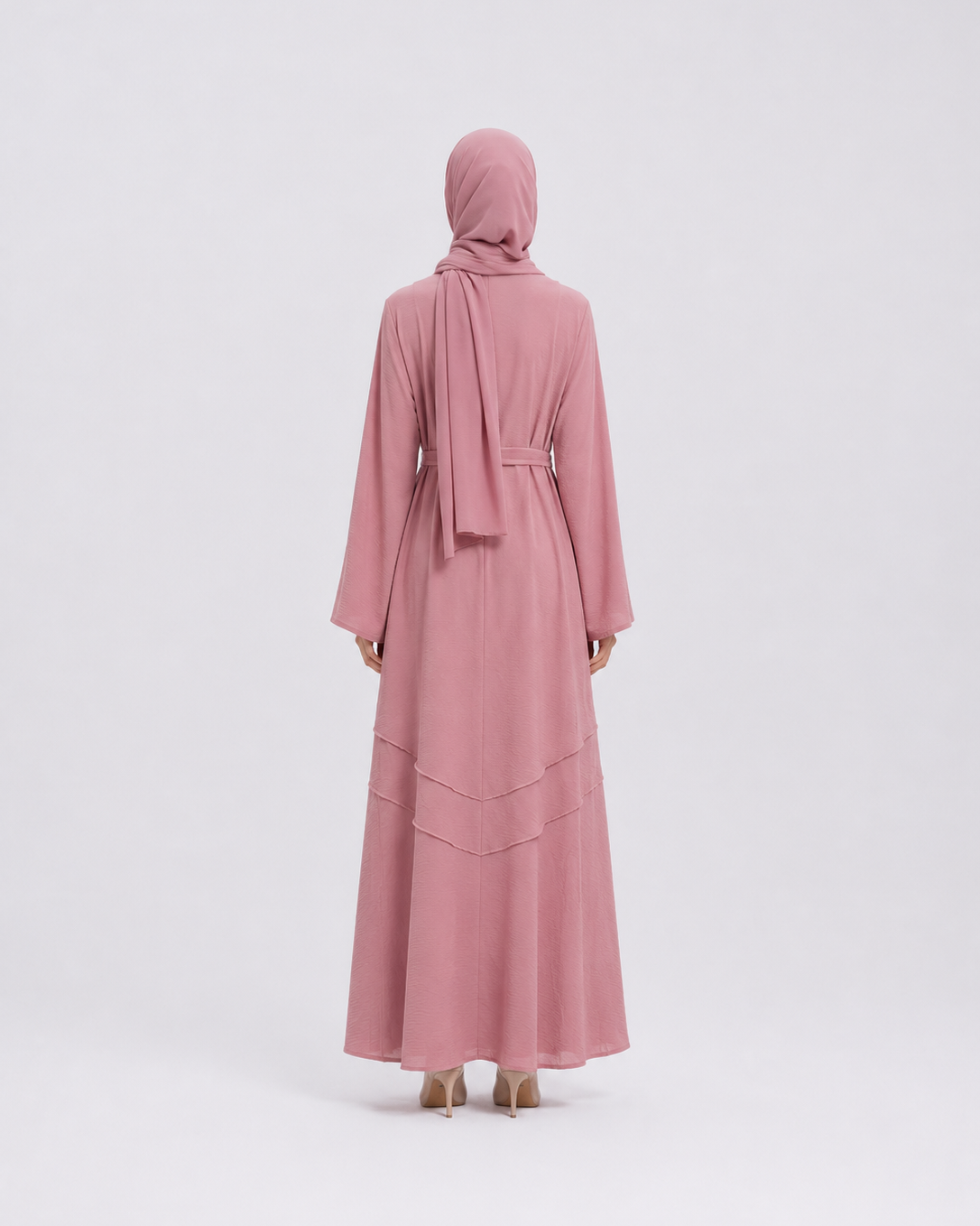A woman viewed from the back wearing a long dusty pink abaya with a matching headscarf and a thin waist tie, standing against a light gray studio background.