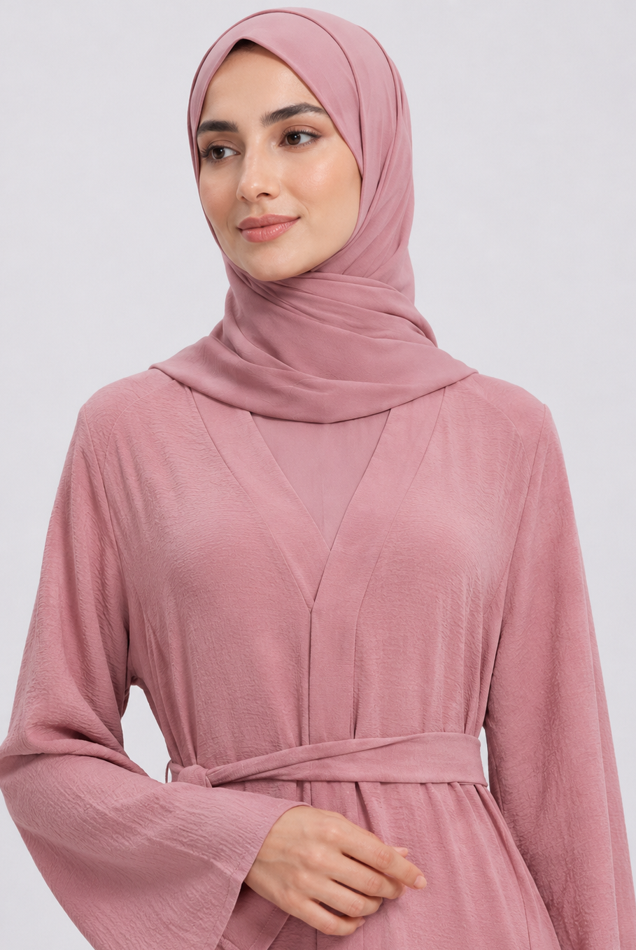 A woman wearing a dusty pink long-sleeve abaya with a matching waist tie belt and a coordinating hijab, photographed against a light gray studio background.