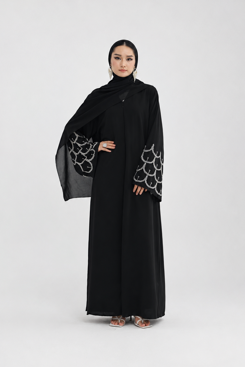 A woman modeling a long black abaya with wide sleeves featuring a white scalloped pattern, paired with a matching black hijab, against a light gray studio background.