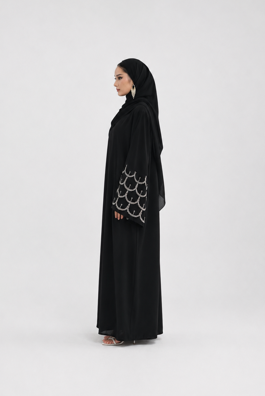 A woman wearing a black abaya with a matching black hijab, standing in profile against a plain light gray studio background. The abaya has a distinctive silver-tone circular pattern on the sleeve.