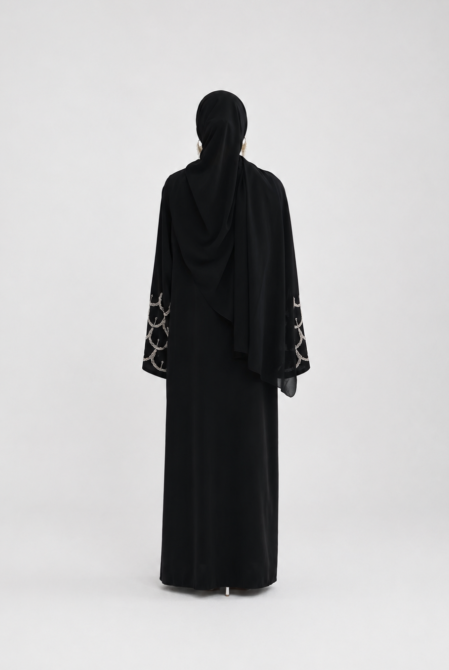 A woman viewed from behind wearing a black abaya with patterned sheer sleeves and a matching black hijab, standing against a plain light gray studio background.