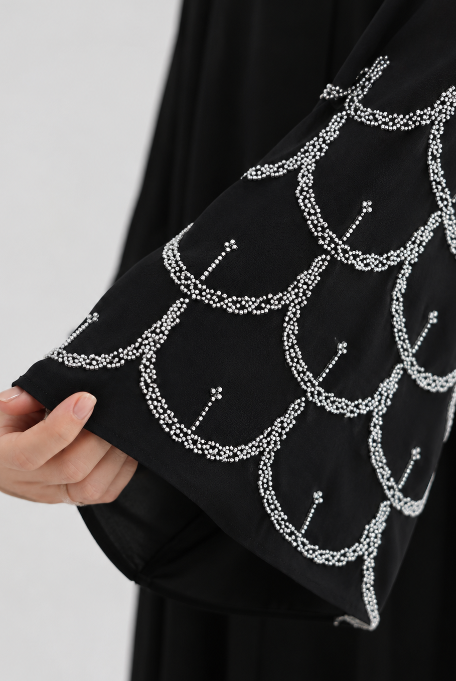 A close-up of a black garment with a wide sleeve decorated with a repeating white scallop or fish-scale pattern made from small dots and lines, held by a hand against a light gray background.