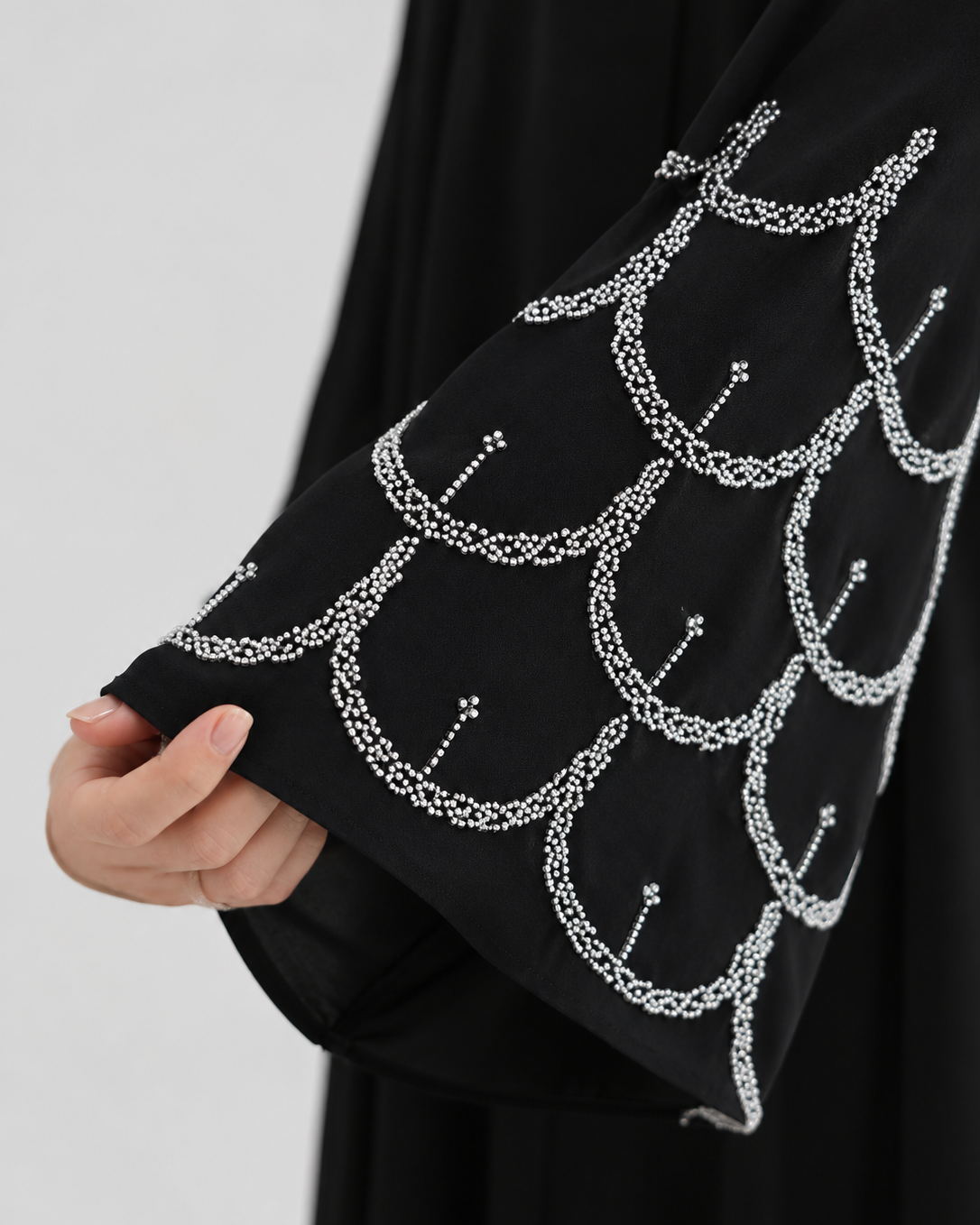 A close-up of a black garment with a wide sleeve decorated with a repeating white scallop or fish-scale pattern made from small dots and lines, held by a hand against a light gray background.