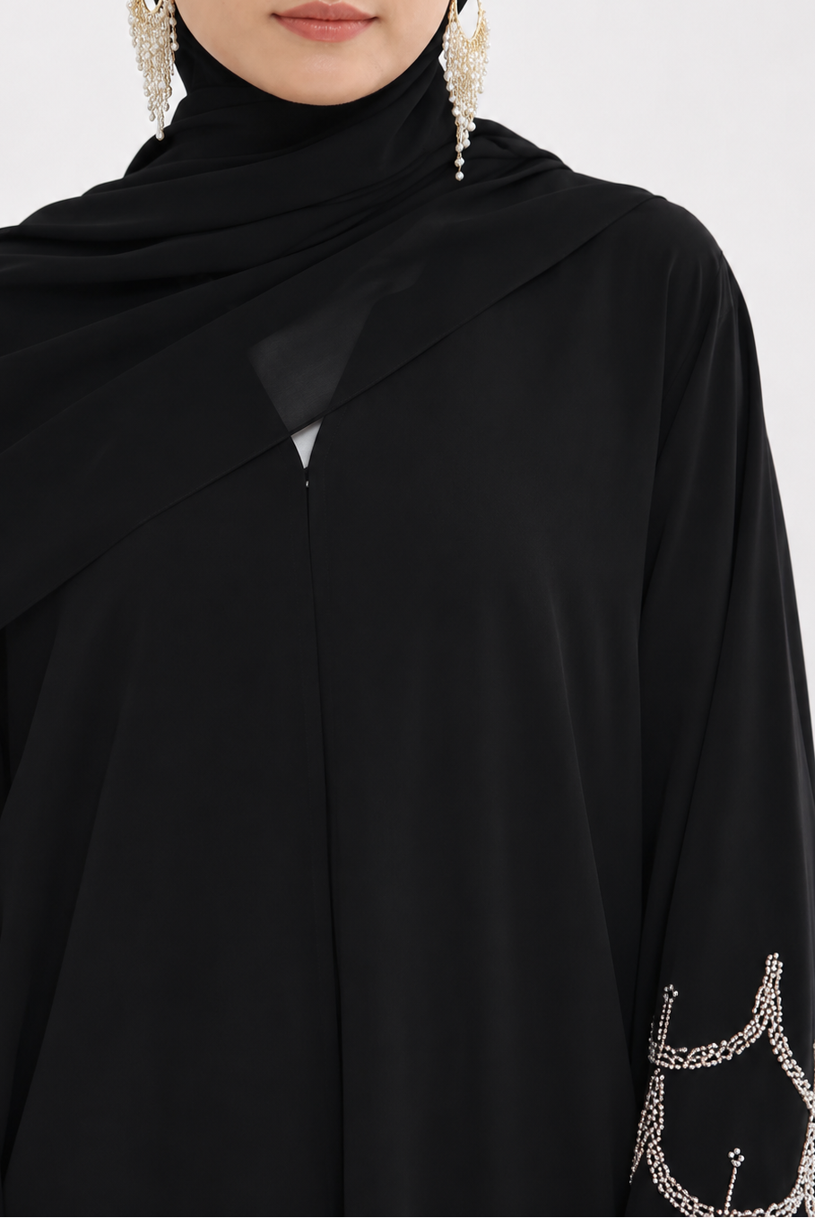 A close-up of a model wearing a black high-neck abaya with draped sleeves and silver chain embroidery on the cuffs, paired with a black hijab and dangling silver earrings.