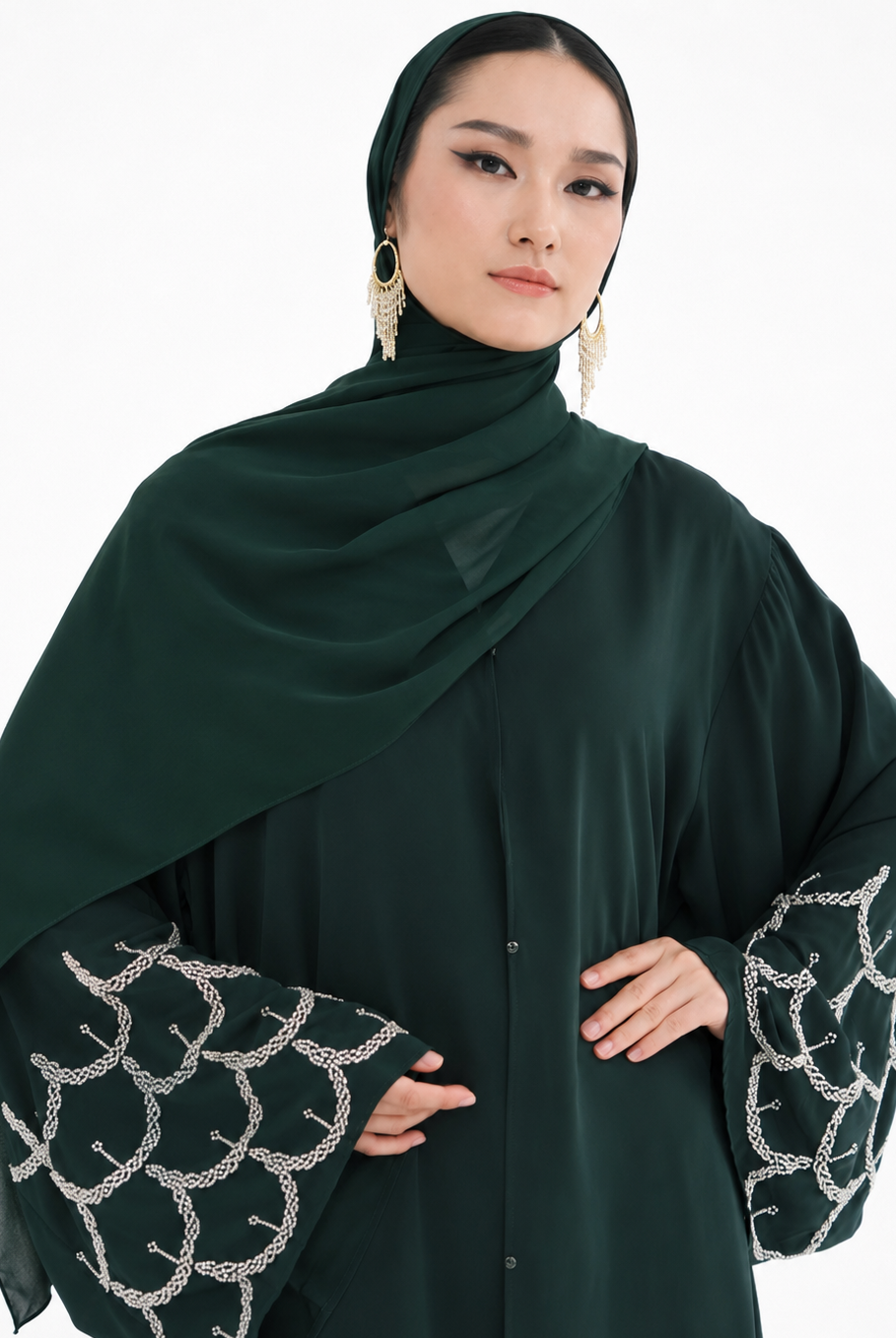 A woman wearing a dark forest green modest outfit featuring a draped hijab, a long-sleeved button-front abaya, and wide bell sleeves with silver chain-link embroidery, set against a plain white background.