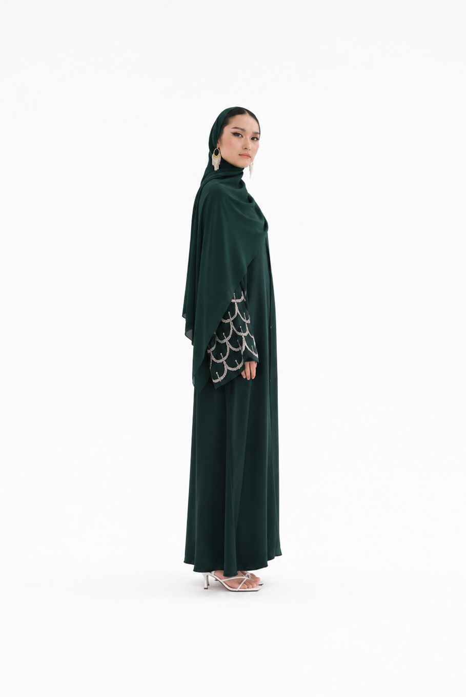 A woman modeling a dark green modest outfit consisting of a long abaya with patterned sleeves and a matching hijab, standing against a plain white studio background.