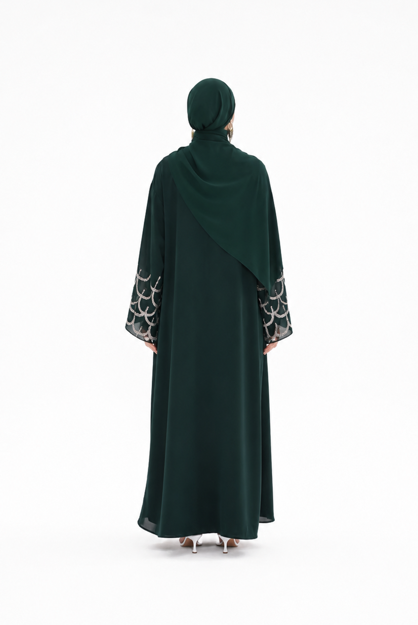 A woman viewed from behind wearing a dark forest green abaya with a matching hijab. The abaya features a draped design with patterned black and white circular lace cuffs on the sleeves.