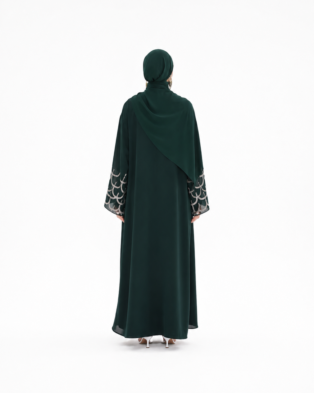 A woman viewed from behind wearing a dark forest green abaya with a matching hijab. The abaya features a draped design with patterned black and white circular lace cuffs on the sleeves.