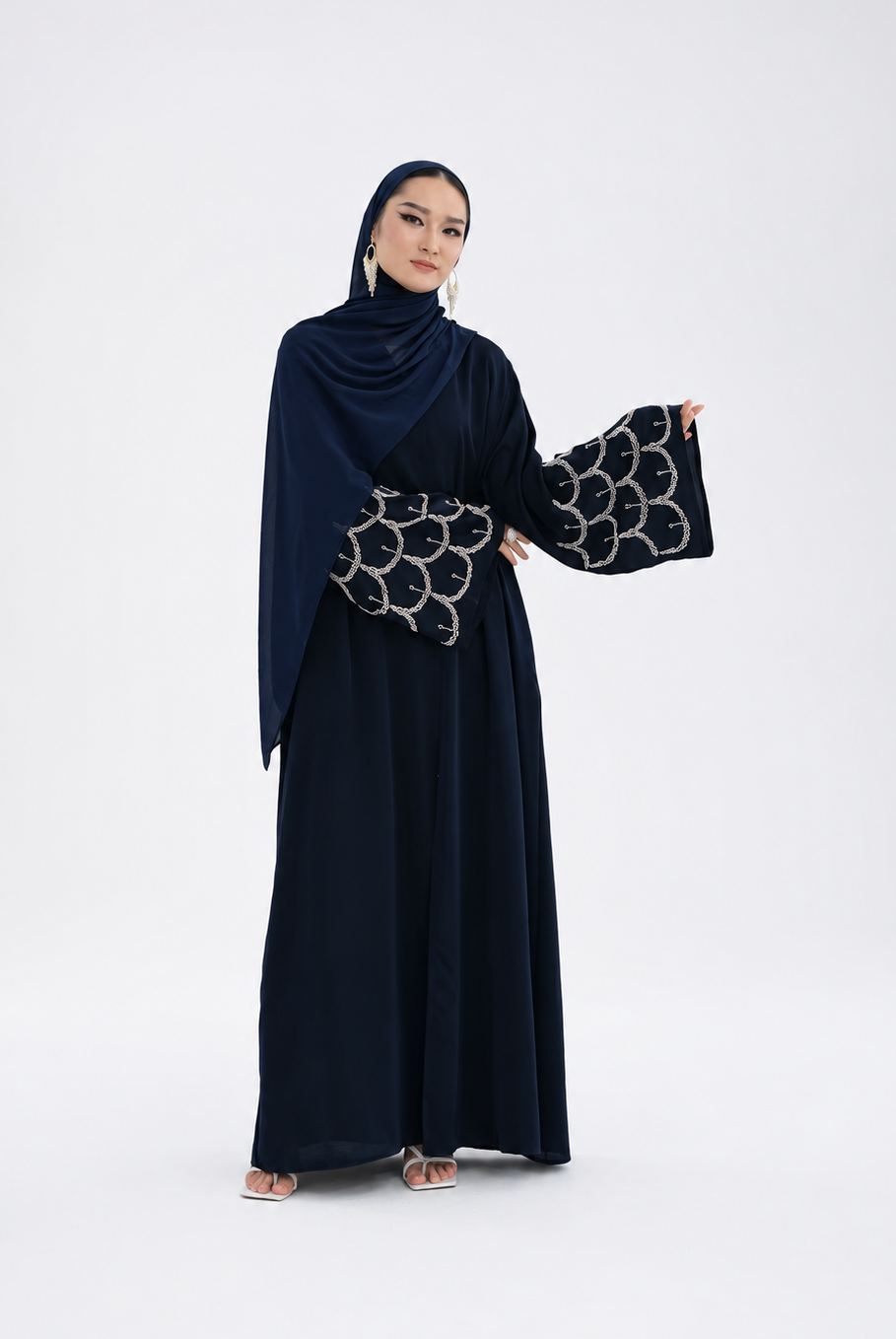 A woman modeling a dark navy blue abaya dress with wide sleeves featuring a silver geometric pattern, paired with a matching draped hijab, against a plain light gray studio background.