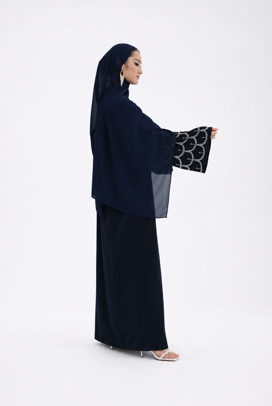 A woman modeling a dark navy abaya with wide sleeves featuring a black and white circular pattern, paired with a matching navy hijab and skirt, set against a plain white studio background.