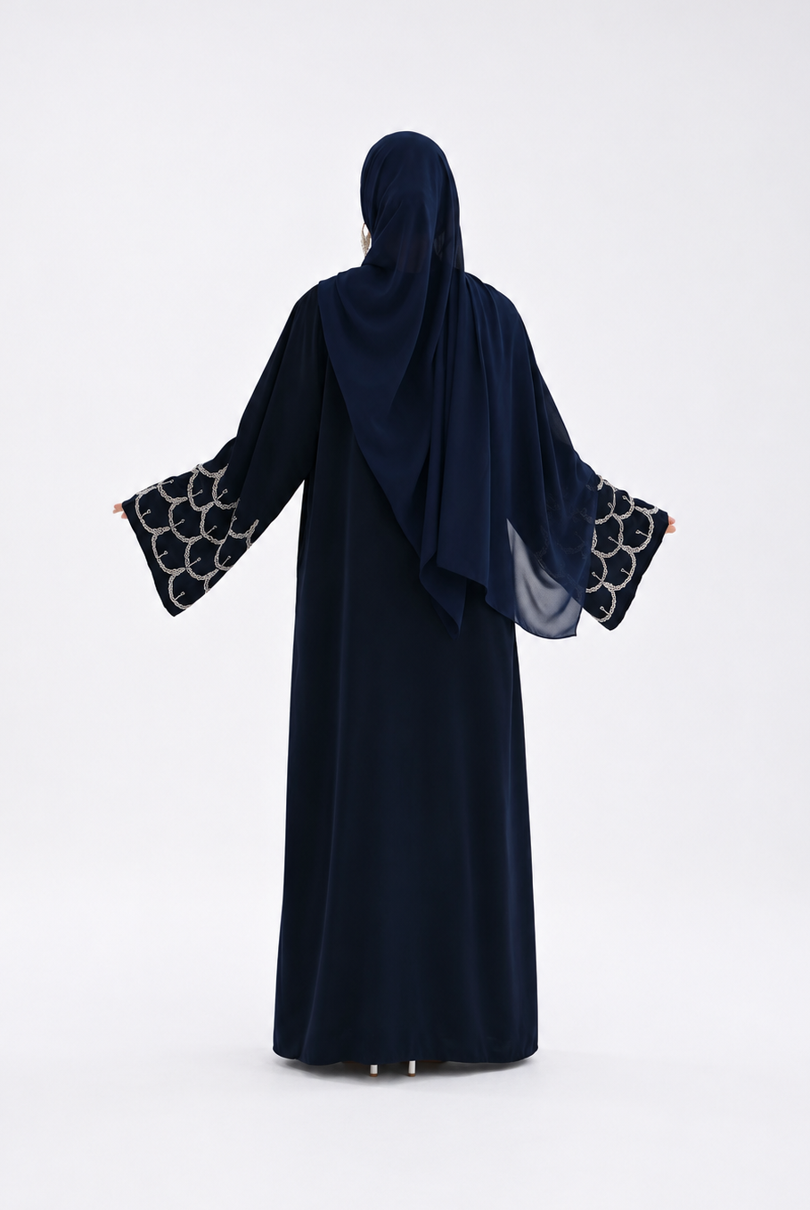 A woman wearing a dark navy abaya and matching hijab, shown from the back against a plain white studio background. The abaya has wide sleeves with a black and white circular patterned trim at the cuffs.