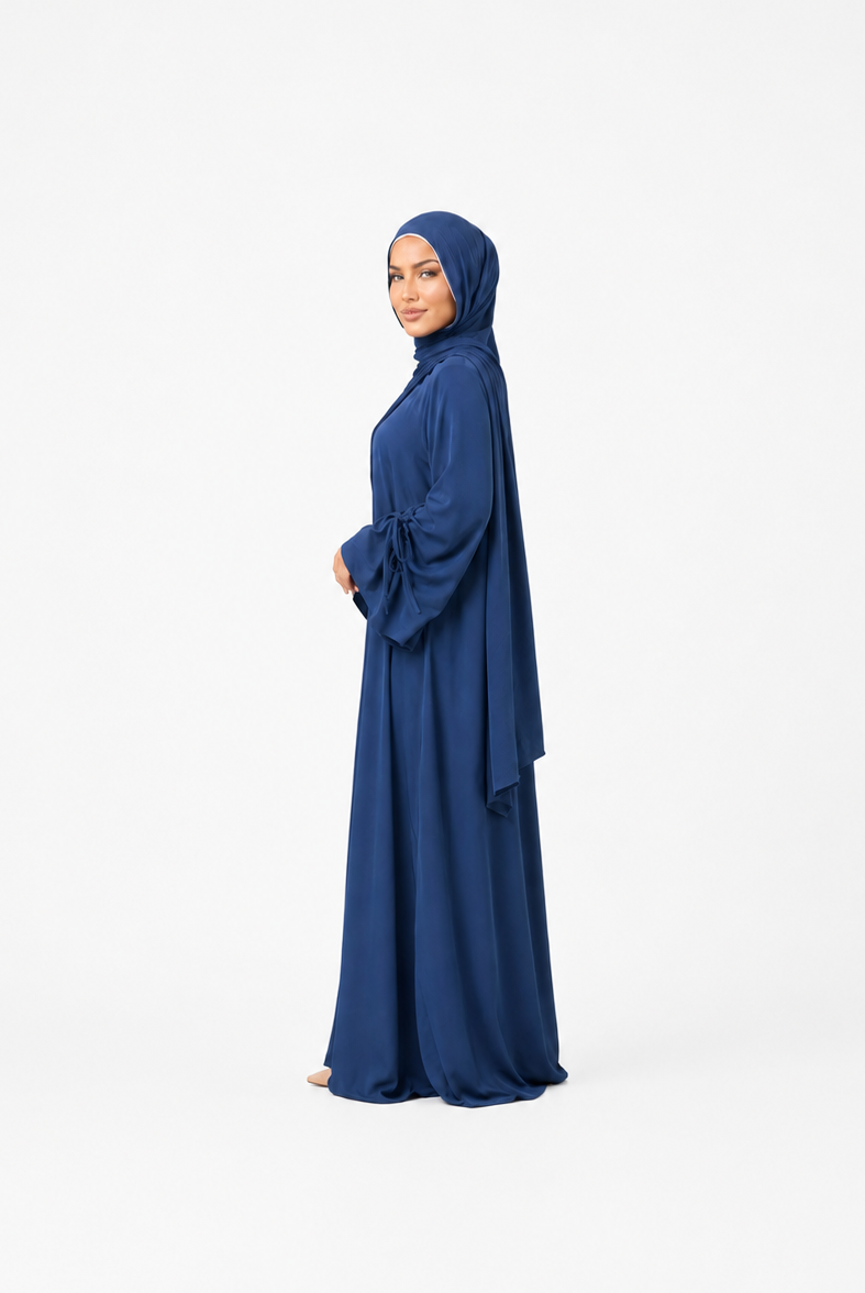 A woman wearing a long navy blue abaya and matching hijab stands in profile against a plain white studio background.