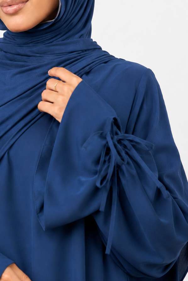 A woman wearing a navy blue modest outfit consisting of a draped hijab and a matching long-sleeve abaya with tie details at the cuffs, photographed against a light gray studio background.