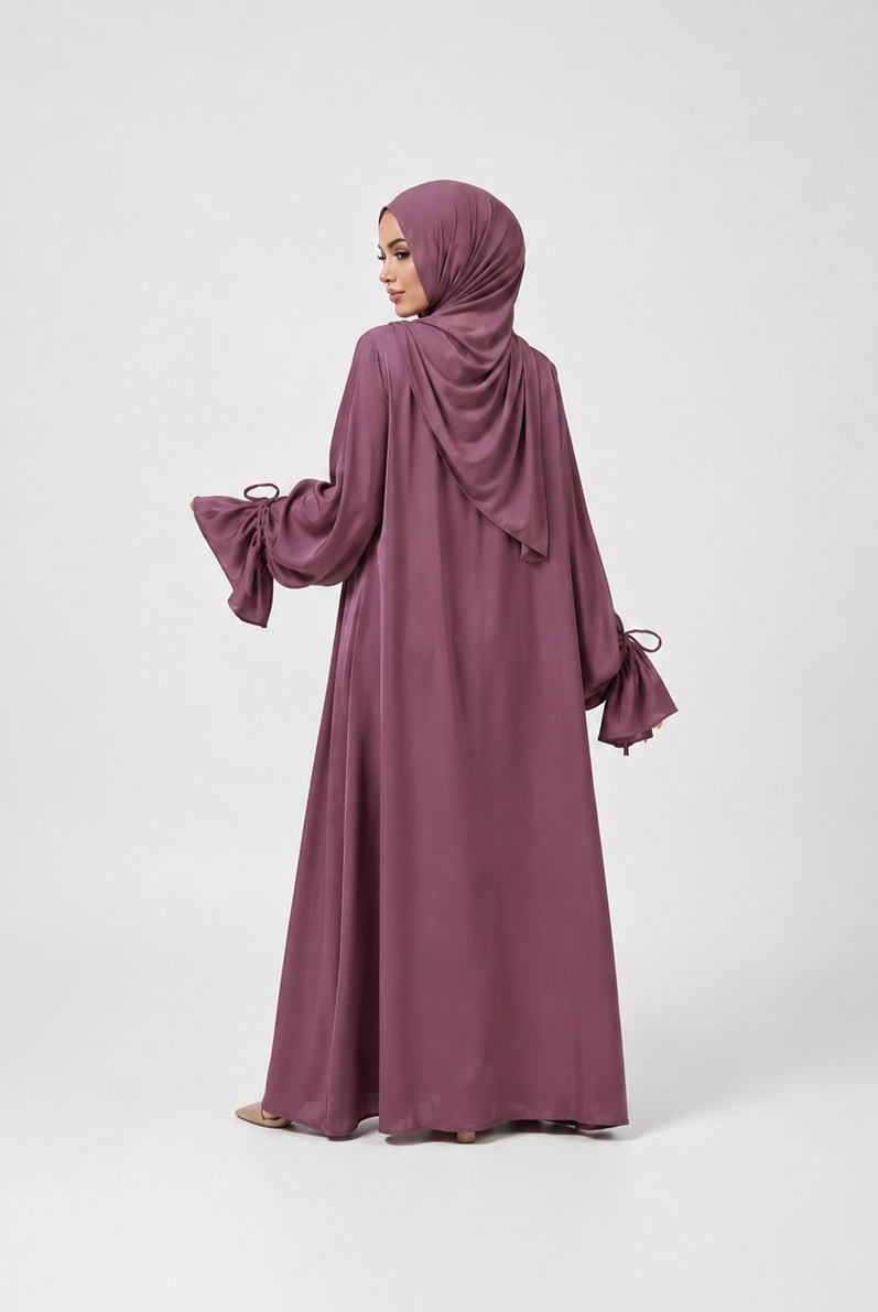 A woman wearing a long, flowing mauve-purple abaya with matching hijab, shown from the back against a light gray studio background.