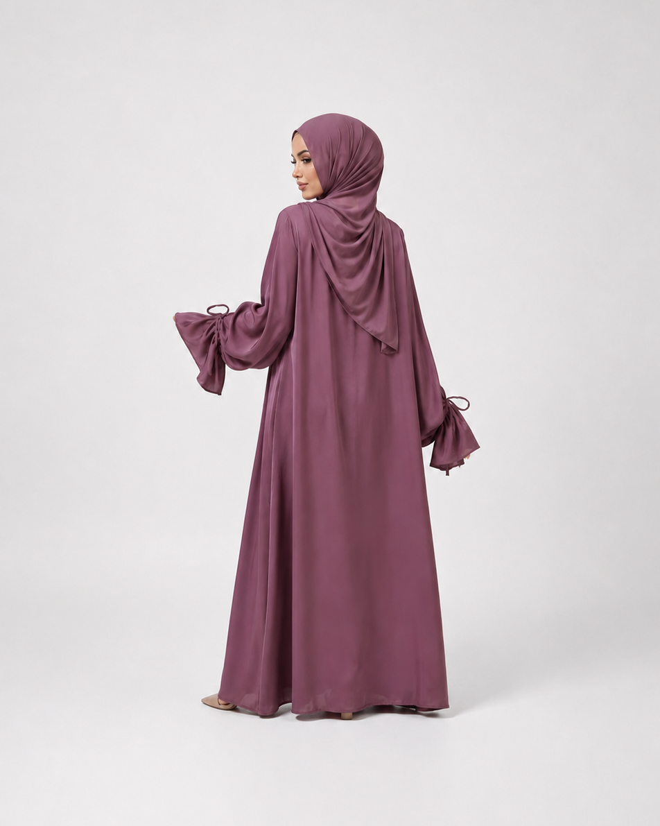 A woman wearing a long, flowing mauve-purple abaya with matching hijab, shown from the back against a light gray studio background.
