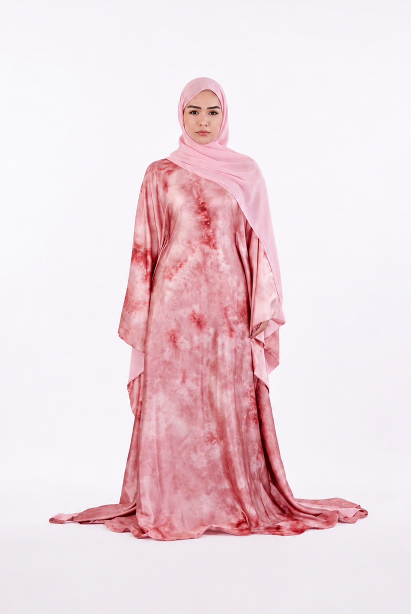 A woman wearing a long pink abaya with a red tie-dye pattern and a matching light pink hijab, standing against a seamless white studio background.