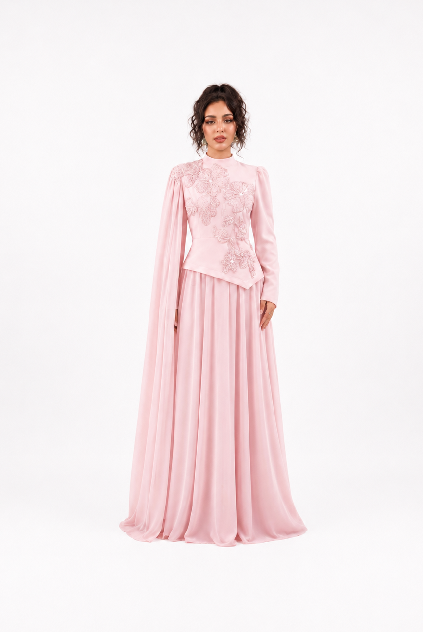 A woman modeling a long pink formal gown with a high neck, sheer draped cape sleeves, and intricate floral embroidery on the bodice, set against a plain white studio background.