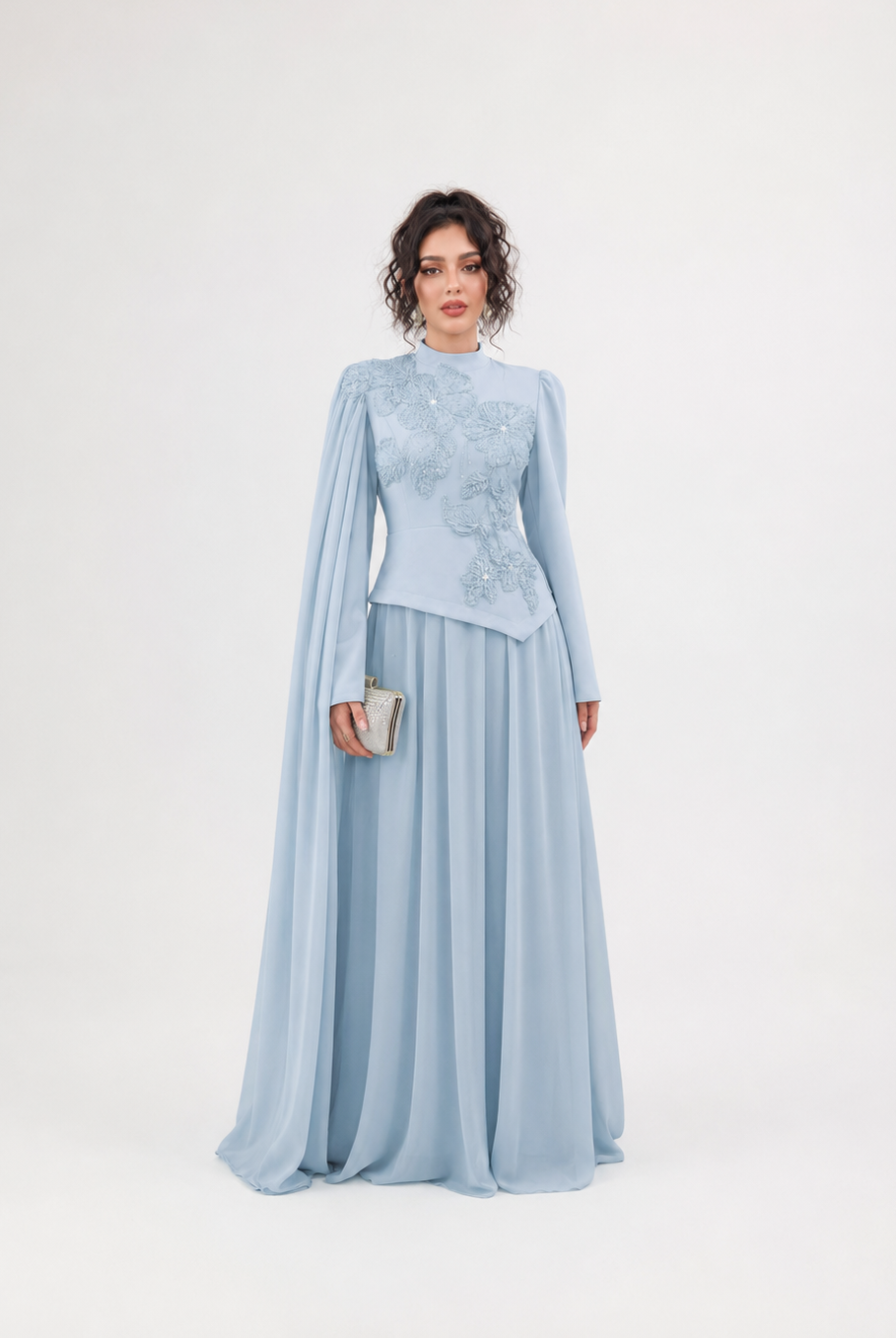 A woman modeling a light blue formal dress with a high mock neck, long draped cape sleeves, and intricate floral embroidery on the bodice, set against a plain white studio background.