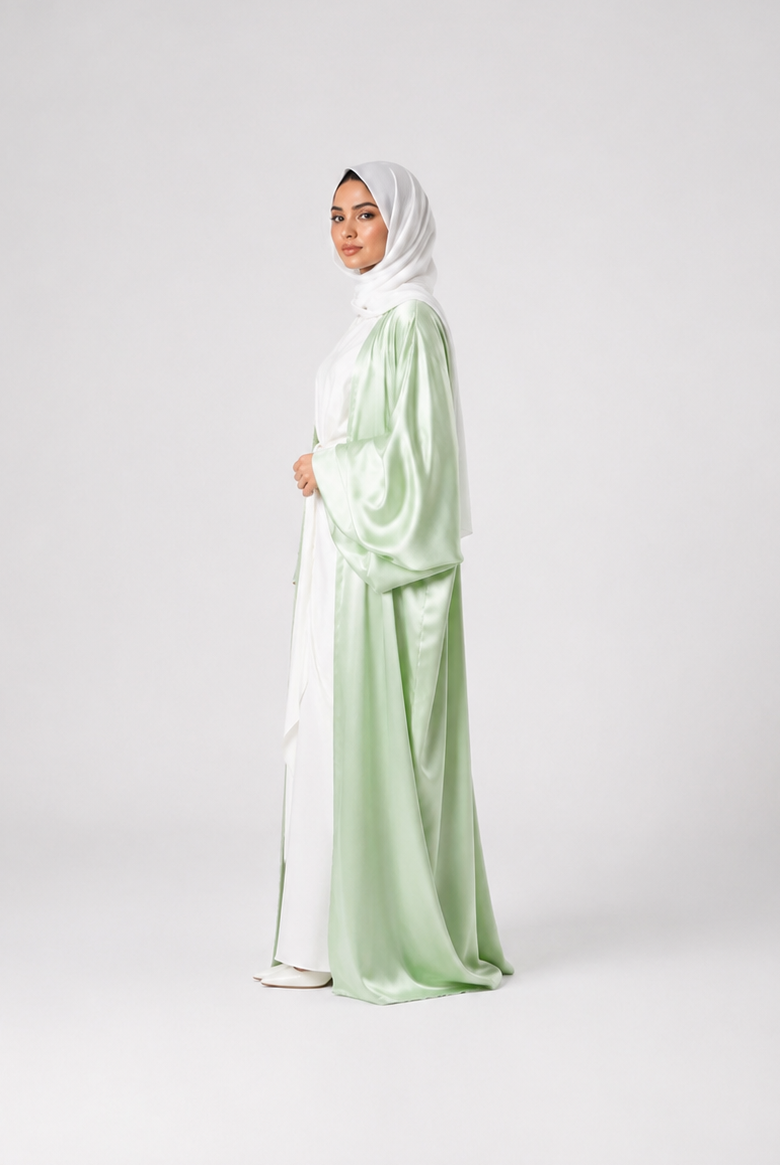 A woman wearing a light sage green and white satin abaya with a matching white hijab, standing in profile against a light gray studio background.