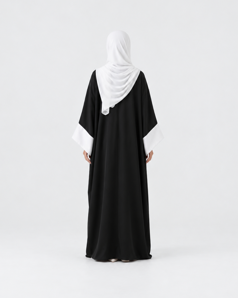 A woman viewed from behind wearing a black abaya with contrasting white sleeve cuffs and a white hijab, standing against a seamless light gray studio background.