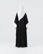A woman viewed from behind wearing a black abaya with contrasting white sleeve cuffs and a white hijab, standing against a seamless light gray studio background.