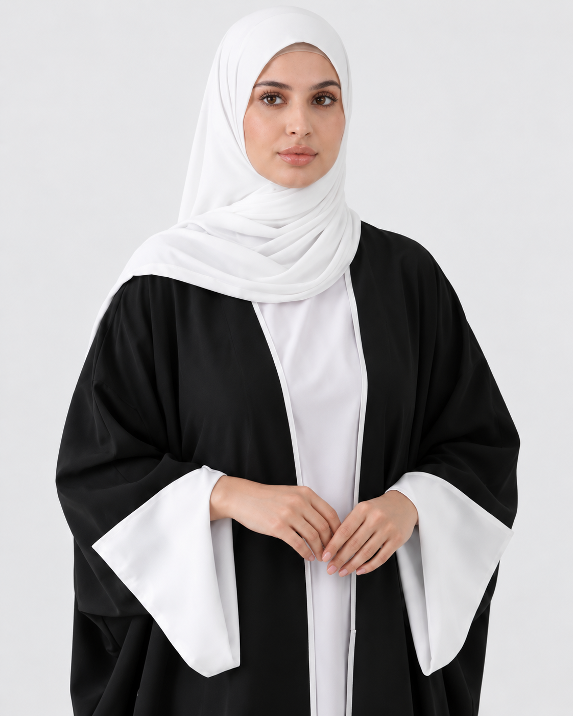 A woman wearing a black abaya with wide white sleeve panels and a white hijab, standing against a light gray studio background.