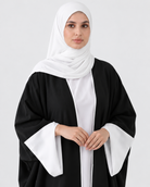 A woman wearing a black abaya with wide white sleeve panels and a white hijab, standing against a light gray studio background.