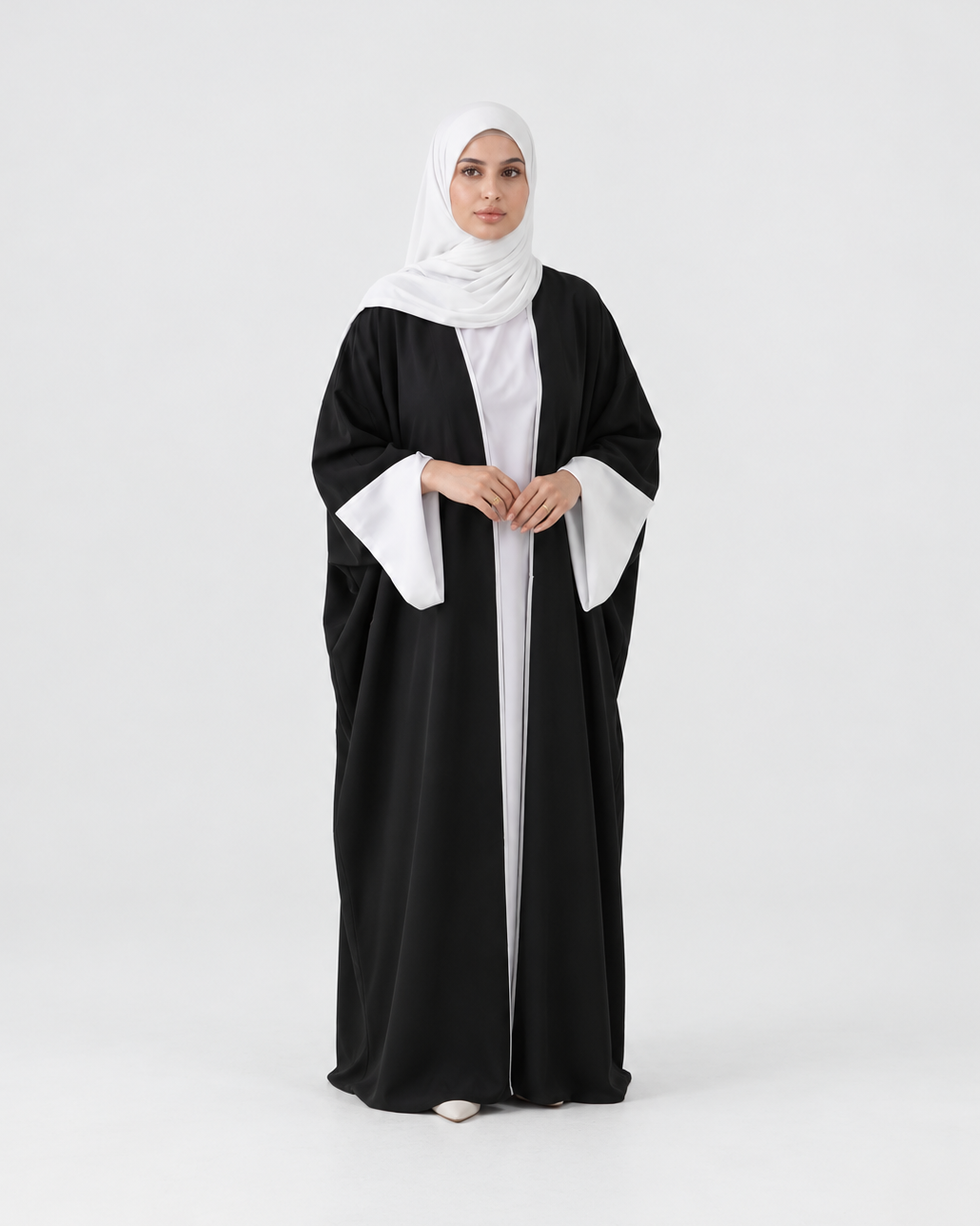 A woman modeling a black abaya with contrasting white wide sleeves, worn over a white inner dress and a white hijab, standing against a light gray studio background.