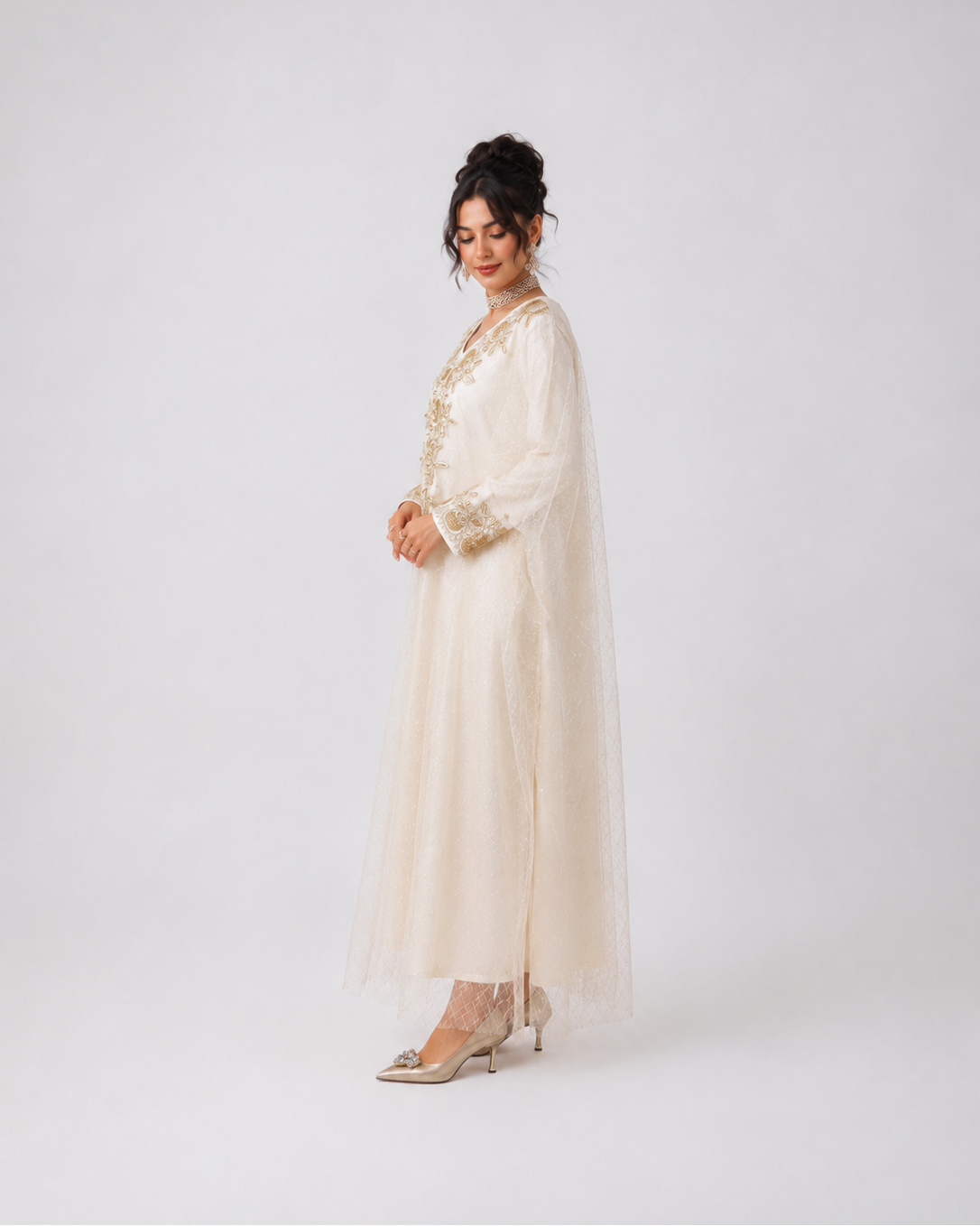 A woman models a long cream-colored tunic dress with sheer sleeves and gold embroidery, paired with a matching sheer veil, against a plain light gray studio background.