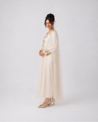 A woman models a long cream-colored tunic dress with sheer sleeves and gold embroidery, paired with a matching sheer veil, against a plain light gray studio background.