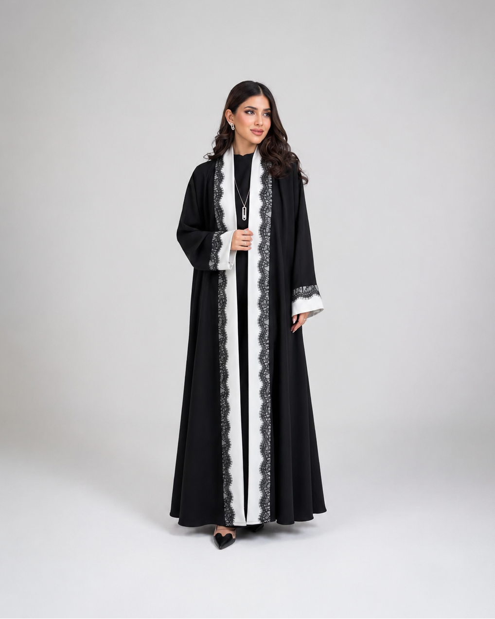 A woman modeling a long black abaya with white lace trim, worn over a black inner dress, against a neutral gray studio background.