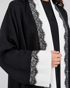 A close-up of a black open-front robe or kimono-style garment with wide sleeves, featuring prominent white vertical panels and black lace trim along the edges and cuffs, worn over a black top.