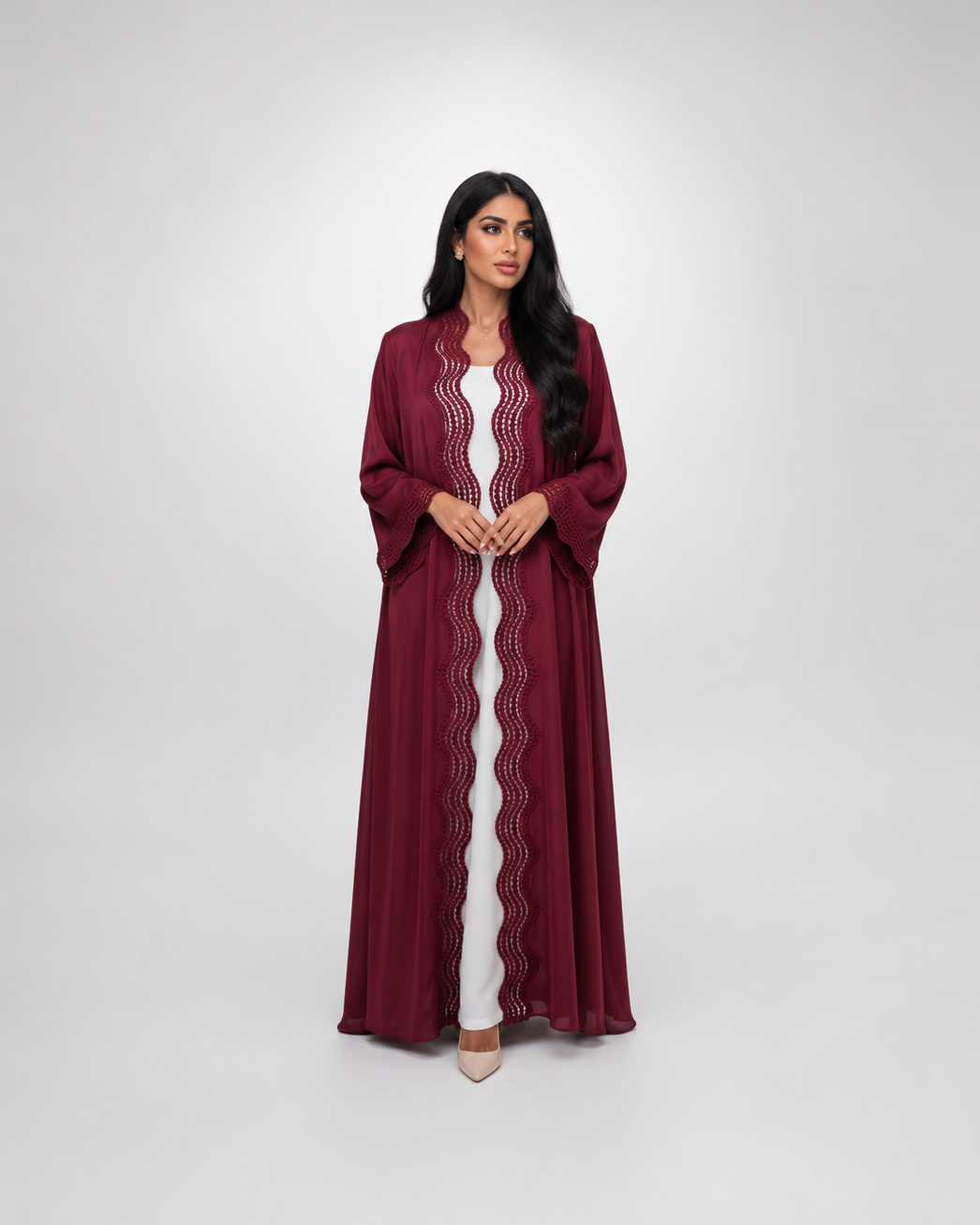 A woman modeling a long, flowing maroon abaya with scalloped lace trim over a white inner dress, standing against a light gray studio background.