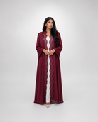 A woman modeling a long, flowing maroon abaya with scalloped lace trim over a white inner dress, standing against a light gray studio background.