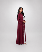 A woman modeling a long, flowing maroon abaya with a matching headscarf, standing against a light gray studio background.