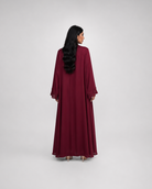 A woman with long black hair is shown from behind wearing a long, flowing maroon abaya with wide sleeves, standing against a light gray studio background.