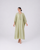 A woman modeling a long, flowing sage green dress with wide sleeves and embroidered detailing on the chest, standing against a light gray studio background.