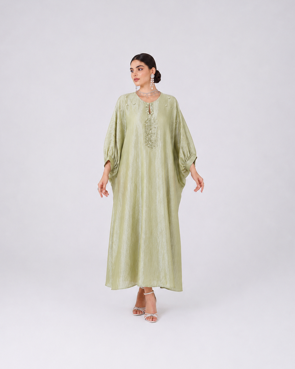 A woman modeling a long, flowing sage green dress with wide sleeves and embroidered detailing on the chest, standing against a light gray studio background.