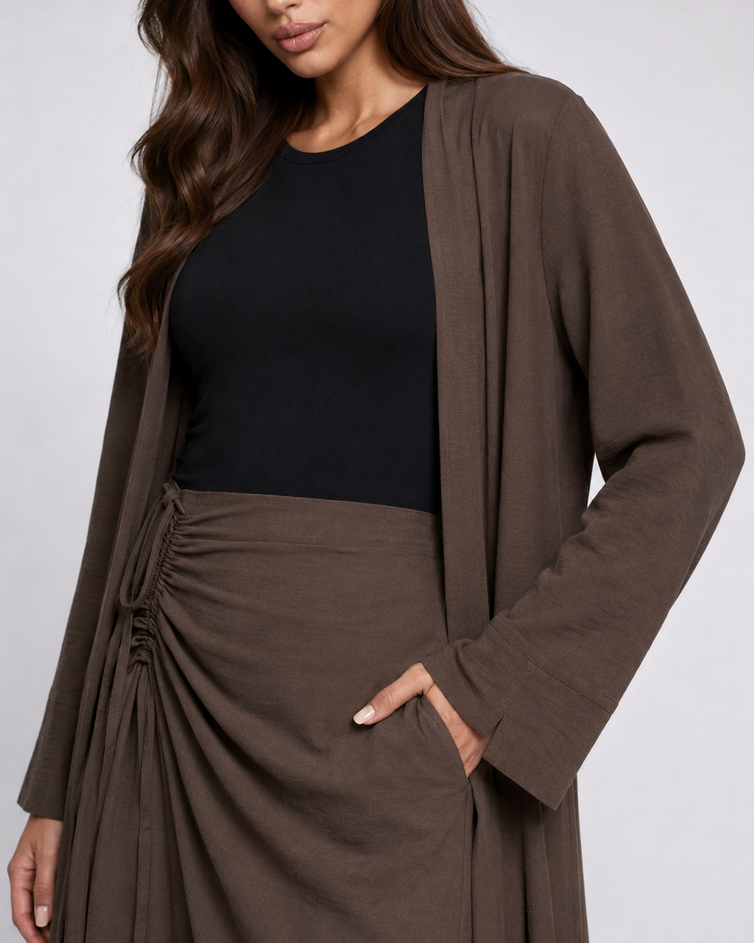 A woman wearing a brown long-sleeve open-front cardigan over a black top and a matching brown ruched skirt, shown against a light gray studio background.