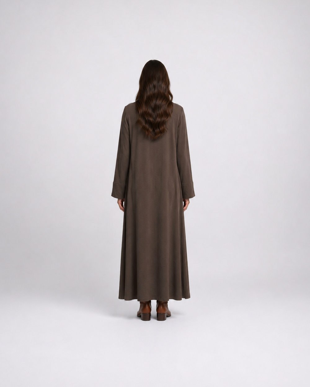 A woman viewed from behind wearing a long, loose-fitting brown maxi dress with wide sleeves, standing against a plain light gray studio background.