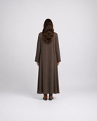 A woman viewed from behind wearing a long, loose-fitting brown maxi dress with wide sleeves, standing against a plain light gray studio background.