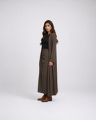 A woman modeling a long brown draped cardigan over a black top and matching brown pleated maxi skirt, standing against a light gray studio background.