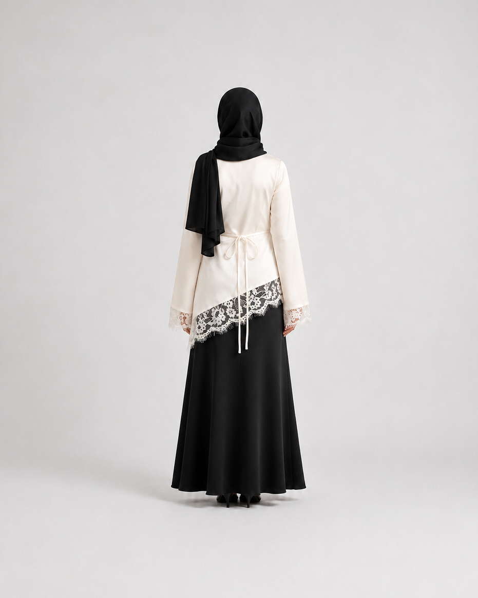 A woman viewed from behind wearing a black hijab, a cream-colored long-sleeve top with black lace trim, and a long black skirt, standing against a light gray studio background.