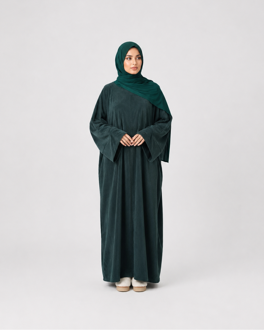 A woman wearing a dark forest green abaya and matching hijab, standing against a light gray studio background.
