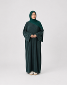 A woman wearing a dark forest green abaya and matching hijab, standing against a light gray studio background.