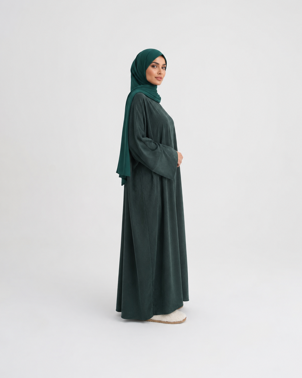 A woman wearing a dark forest green abaya and matching hijab stands in profile against a seamless light gray studio background.