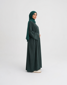 A woman wearing a dark forest green abaya and matching hijab stands in profile against a seamless light gray studio background.